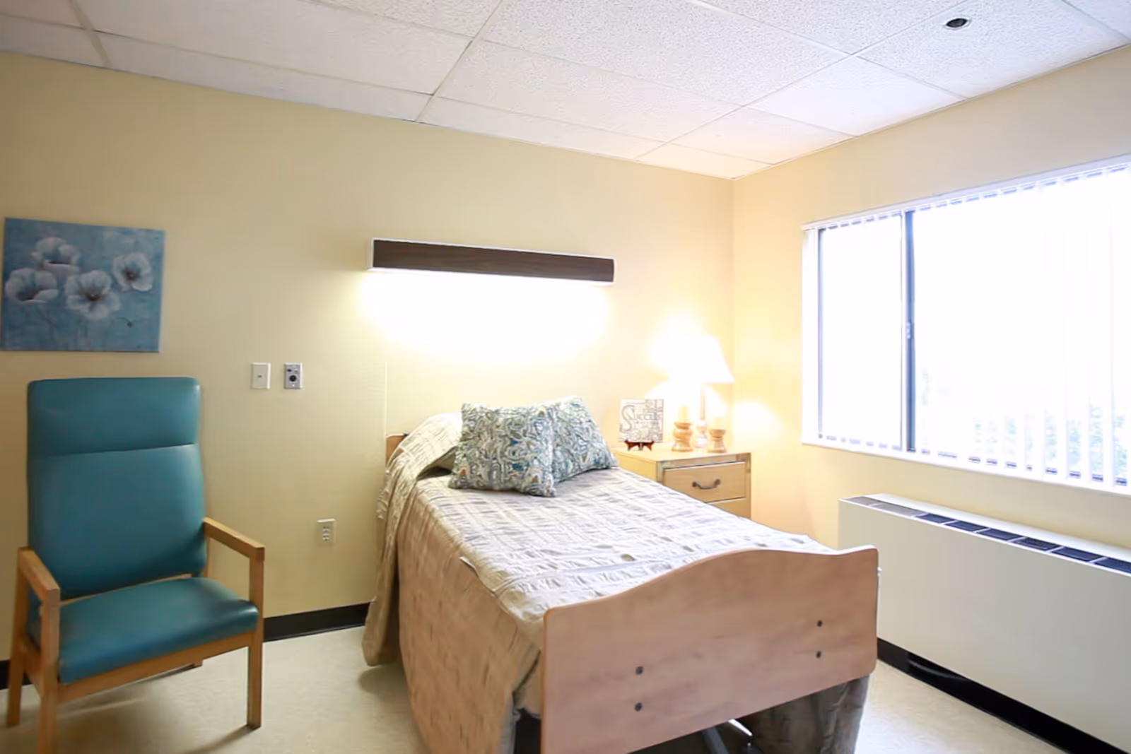 Small furnished resident bedroom with a single bed, bedside table and lamp, teal armchair, and a window with vertical blinds.