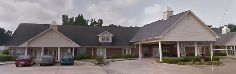 Front exterior of a single-story senior living building with a covered porte-cochere, parked cars, and a small fountain in the driveway.