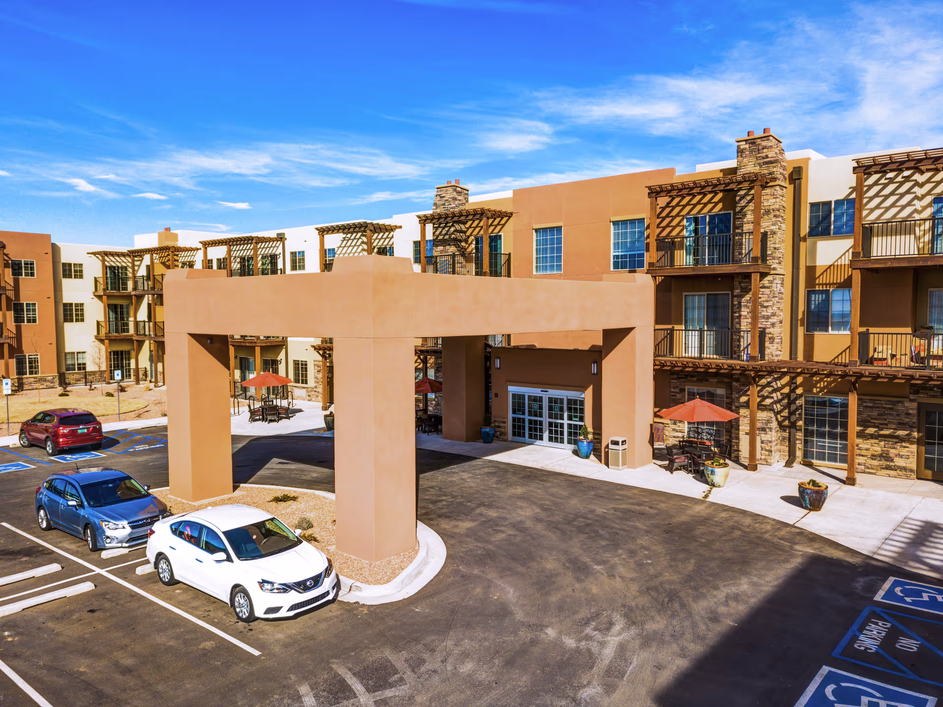 Exterior view of a senior living facility with a large covered entrance, multiple balconies, stone and stucco facade, outdoor seating with umbrellas, and a parking lot with several cars and handicap parking spaces.