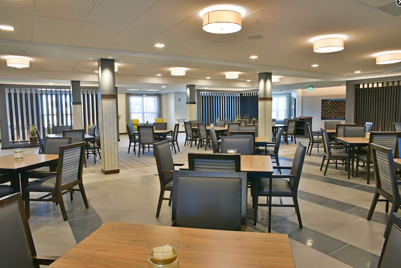 A spacious dining room with multiple wooden tables and black cushioned chairs arranged neatly. The room features several pillars with tile accents, large windows with vertical blinds, and modern ceiling lights providing warm illumination.