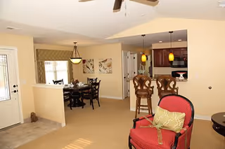Interior view of a senior living facility showing a living area with a red armchair and a green pillow, a kitchen with two bar stools and pendant lights, and a dining area with a round table and chairs. The walls are painted beige and there is a ceiling fan above.