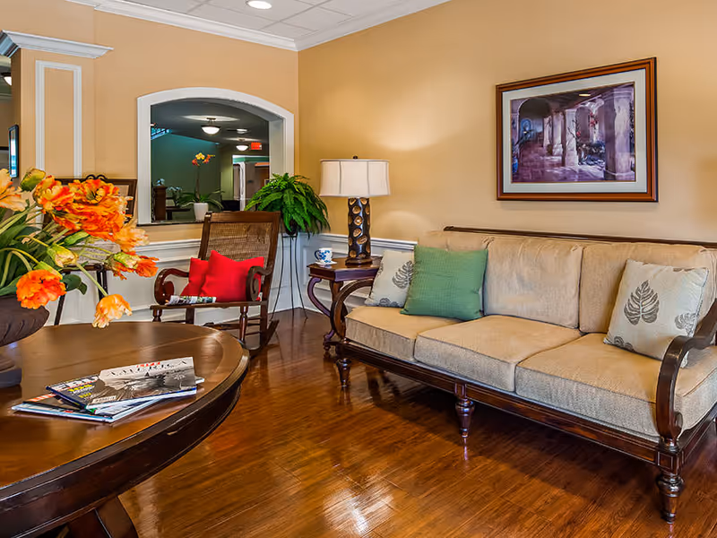 A cozy living room area with a beige sofa adorned with green and patterned pillows, a wooden rocking chair with a red cushion, a side table with a lamp, a round wooden coffee table with magazines, and a vase of orange flowers. The walls are painted light yellow with white trim, and a framed artwork hangs above the sofa. A large mirror reflects part of the adjacent hallway.