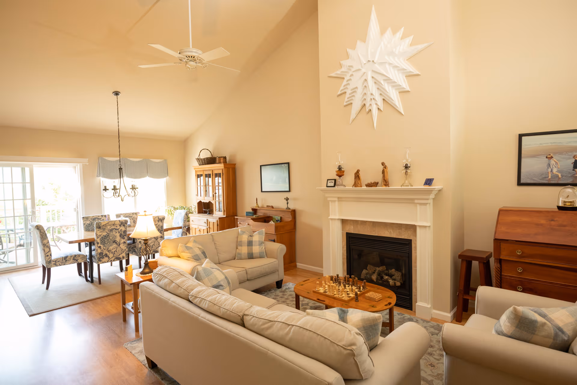 A bright and cozy living room with beige walls and high ceilings featuring a white ceiling fan. The room includes two beige sofas with plaid pillows, a wooden coffee table with a chess set, a fireplace with decorative items on the mantel, and a wooden cabinet. In the background, there is a dining area with a wooden table and floral upholstered chairs near a sliding glass door letting in natural light.