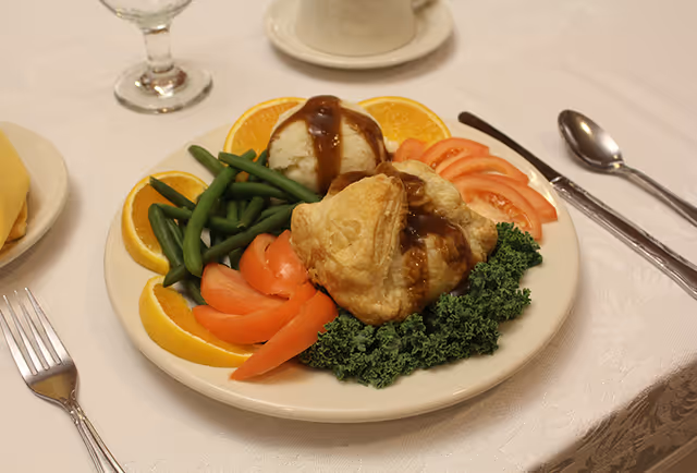 A plate of food featuring a puff pastry with gravy, mashed potatoes topped with gravy, green beans, sliced tomatoes, kale, and orange slices, set on a white tablecloth with a fork, knife, spoon, water glass, and coffee cup nearby.