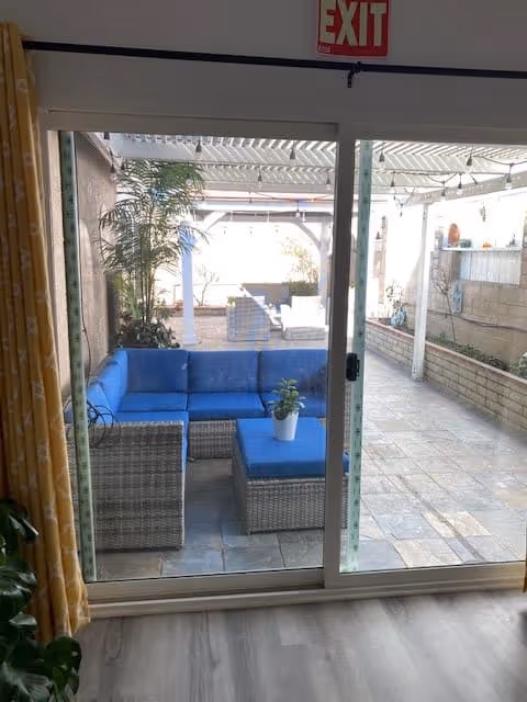 View through a sliding glass door showing an outdoor patio area with a wicker sectional sofa with blue cushions and a matching ottoman with a small potted plant on top. The patio is covered with a pergola and has string lights hanging. There are plants along the left side and a brick planter on the right side.
