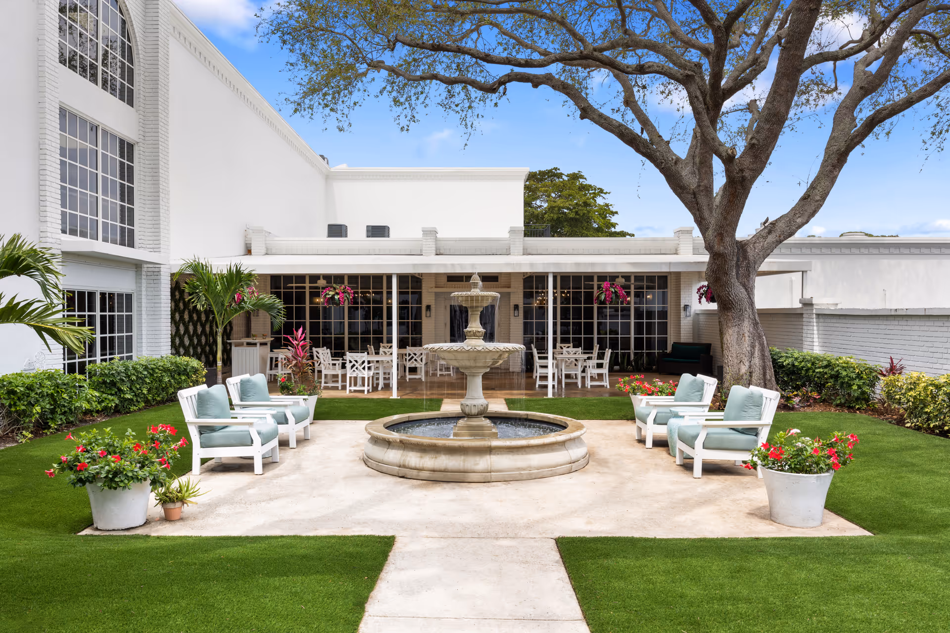 A bright courtyard featuring a central tiered fountain, cushioned patio chairs, potted flowers and a large tree in front of a white building.
