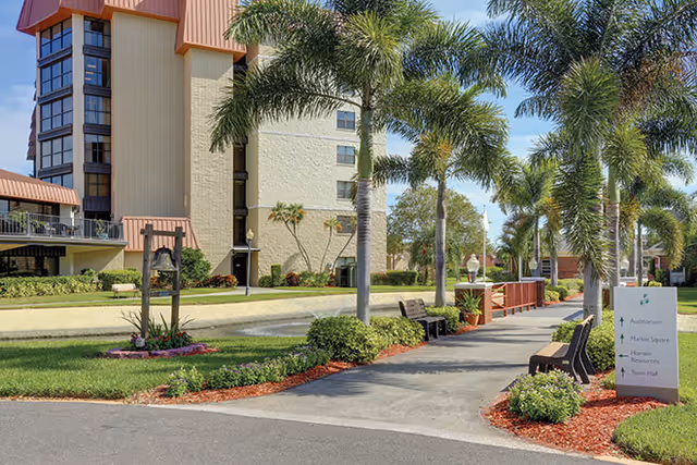 Outdoor walkway lined with palm trees and benches leading to a multi-story building with beige and brown exterior. There are landscaped areas with grass, shrubs, and mulch. A signpost on the right side of the walkway points to Auditorium, Market Square, Human Resources, and Town Hall.