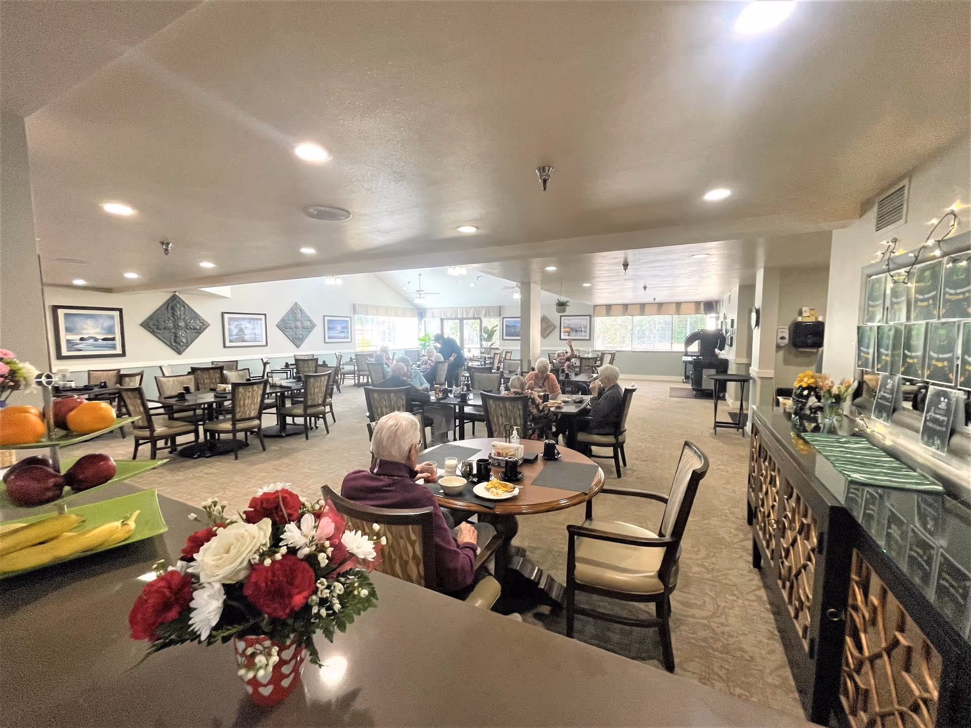 A dining room in a senior living facility with several elderly residents seated at round tables eating and socializing. The room is well-lit with ceiling lights and large windows letting in natural light. There are decorative wall hangings and framed pictures on the walls. A counter with a vase of flowers and a tiered fruit stand is visible in the foreground.