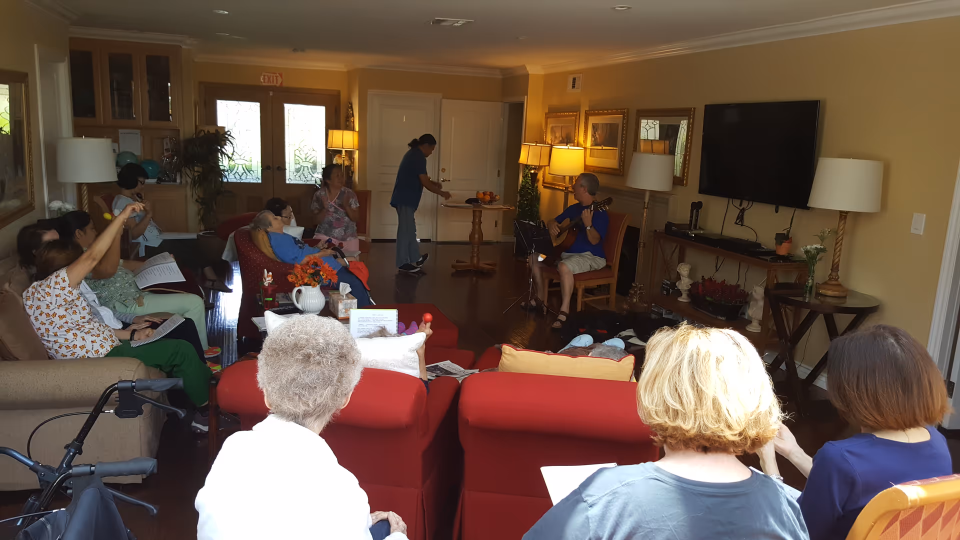 A group of elderly people seated in a living room with red armchairs and beige walls, listening to a man playing guitar. A caregiver is standing near the back door, and the room is warmly lit with lamps and natural light coming through glass doors.