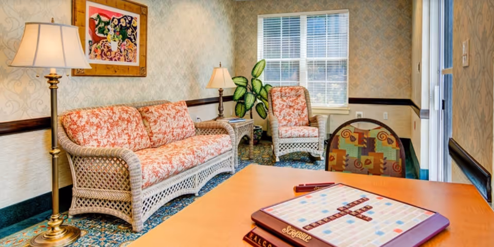 A cozy living room area with a wicker sofa and armchair featuring floral cushions, a floor lamp, a side table with a lamp, a leafy green plant near a window with blinds, and a table in the foreground with a Scrabble board game set up.