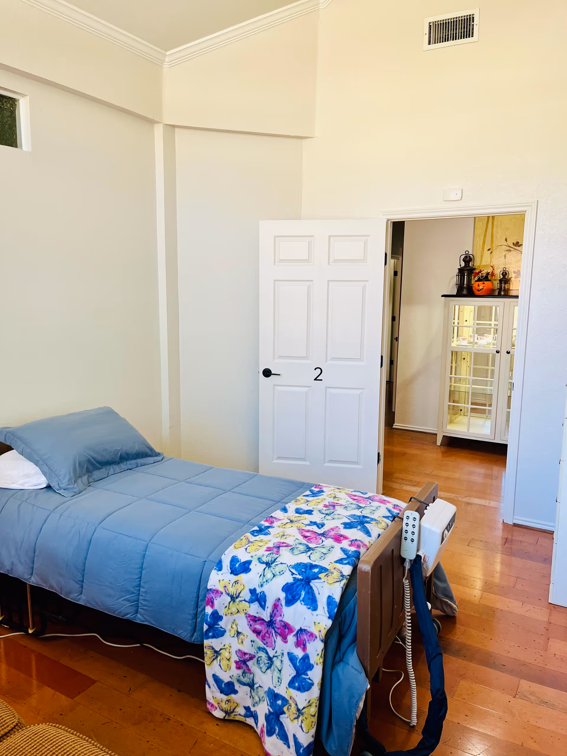 A simple assisted-living bedroom with a single bed in blue bedding and a colorful butterfly throw, a partially open door showing a hallway cabinet.