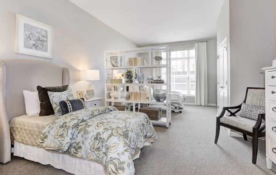 A bright and spacious bedroom with a large window letting in natural light. The room features a bed with floral bedding and multiple pillows, a white nightstand with a lamp and clock, a white open shelving unit with decorative items and books, a cushioned chair with a patterned pillow, and a white dresser. The walls are painted light gray and the carpet is a neutral tone.