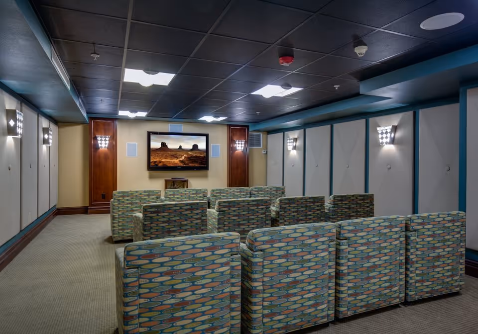 A small theater room with multiple rows of patterned armchairs facing a wall-mounted flat screen TV displaying a desert landscape. The room has a dark ceiling with recessed lighting and wall sconces providing soft illumination.