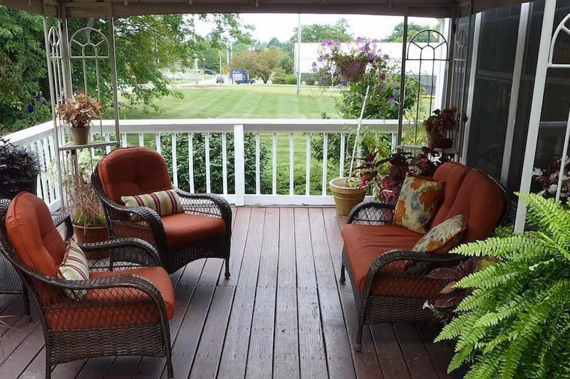 Covered wooden porch with wicker chairs and a loveseat with red cushions, potted plants, and a view of a grassy lawn.