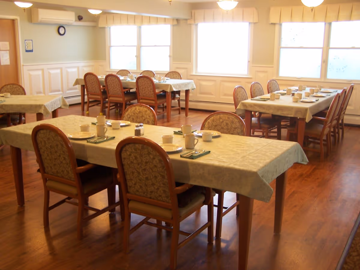 A dining room with several tables covered with light-colored tablecloths, each set with plates, cups, utensils, and napkins. The room has wooden floors, patterned cushioned chairs, large windows with valances, and soft lighting from ceiling fixtures.