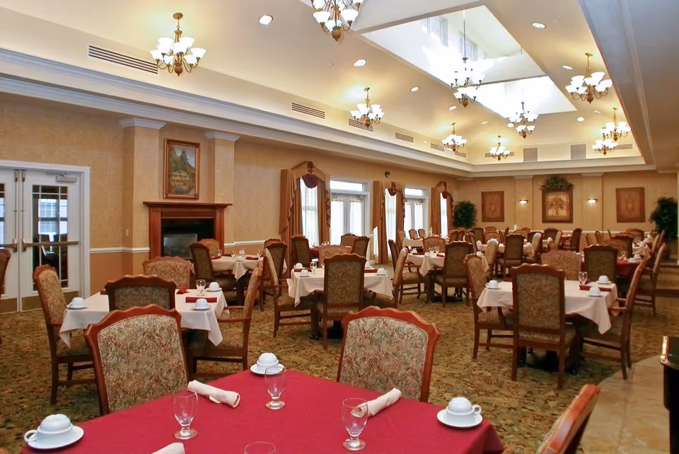 A spacious dining room with multiple tables covered in white and red tablecloths, each set with cups, glasses, and napkins. The room features ornate chairs with patterned upholstery, a fireplace with a framed painting above it, large windows with draped curtains, and multiple chandeliers hanging from a high ceiling with skylights.