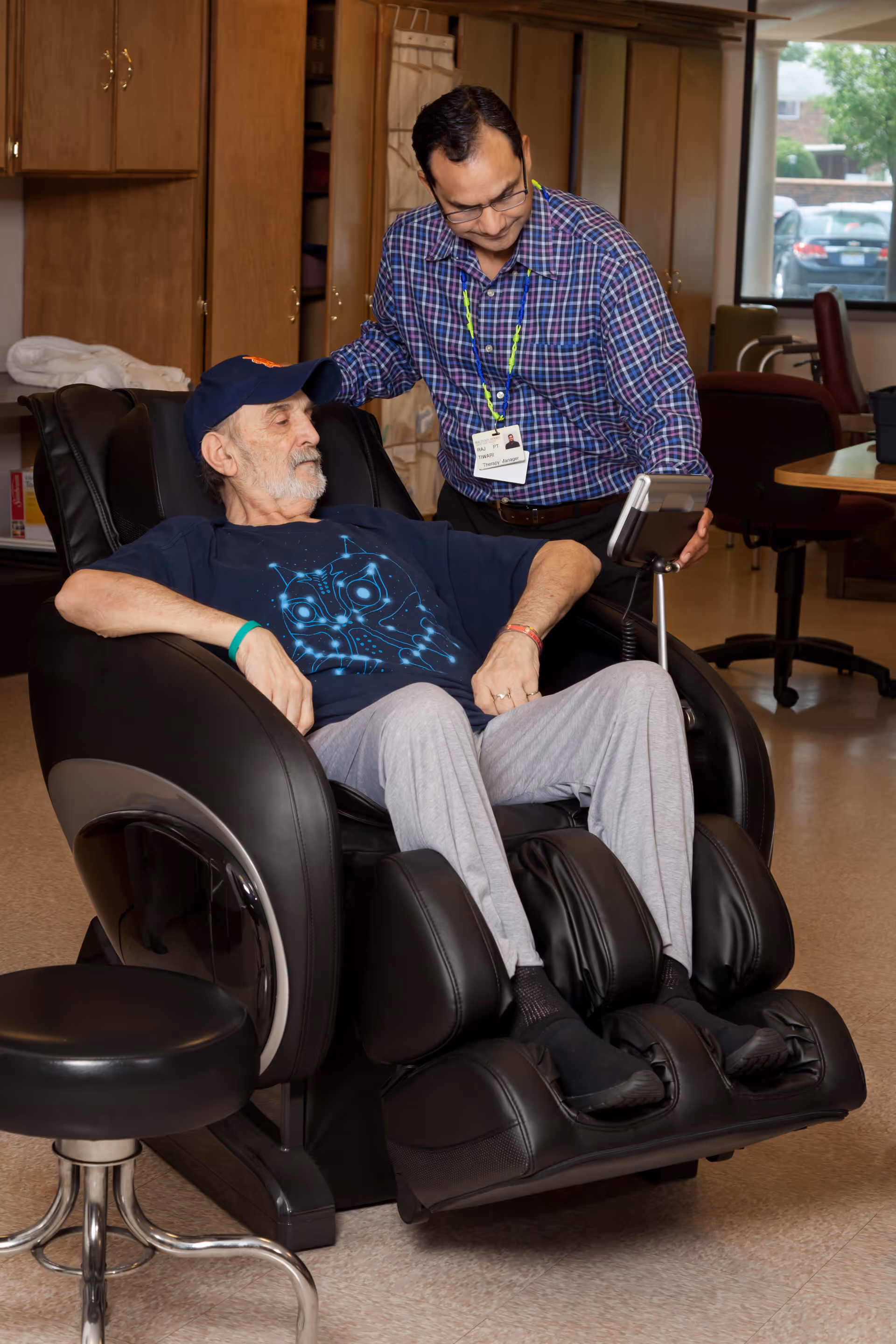 An elderly man wearing a blue graphic t-shirt and gray pants sits comfortably in a black massage chair while a healthcare worker in a plaid shirt and glasses stands beside him, adjusting the chair's control panel in a room with wooden cabinets and office furniture.