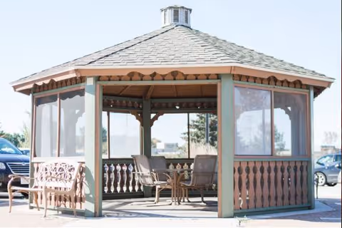 A wooden gazebo with a shingled roof and open sides, featuring a small table and chairs inside. There is a wooden bench outside the gazebo, and cars are parked in the background under a clear sky.