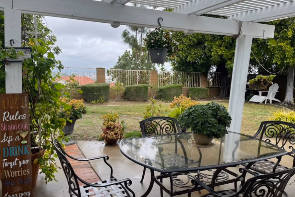 Covered patio with a glass-top dining table, wrought-iron chairs and potted plants overlooking a grassy yard and fence.