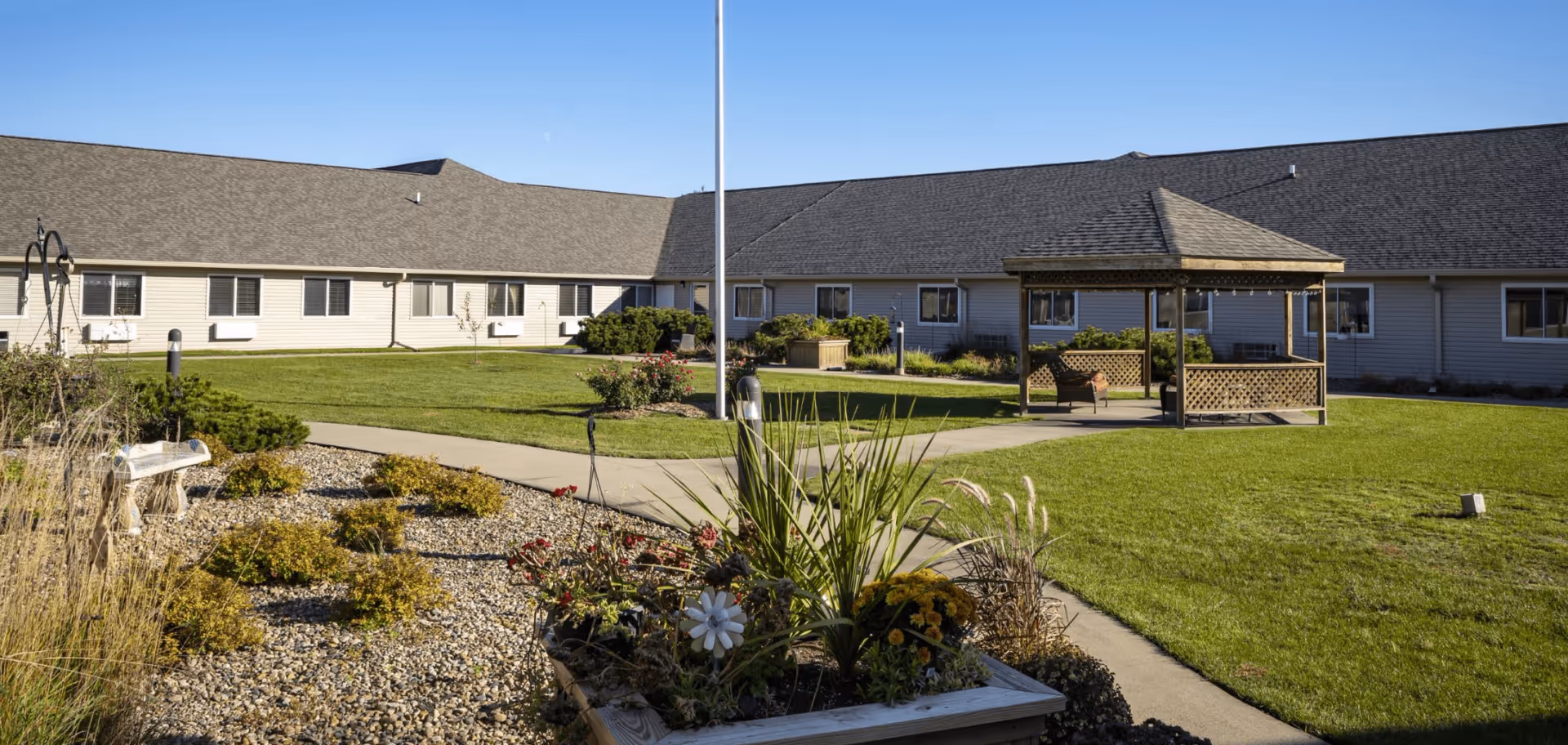 Sunlit courtyard with a gazebo, flagpole, flowerbeds and a surrounding single-story assisted living building.