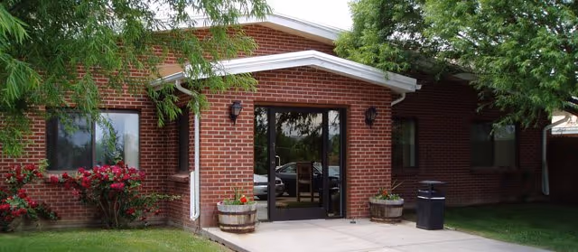 Entrance of a single-story brick building with glass double doors, flanked by two wooden barrels with flowers. There are bushes with red flowers on the left and trees providing shade around the building.