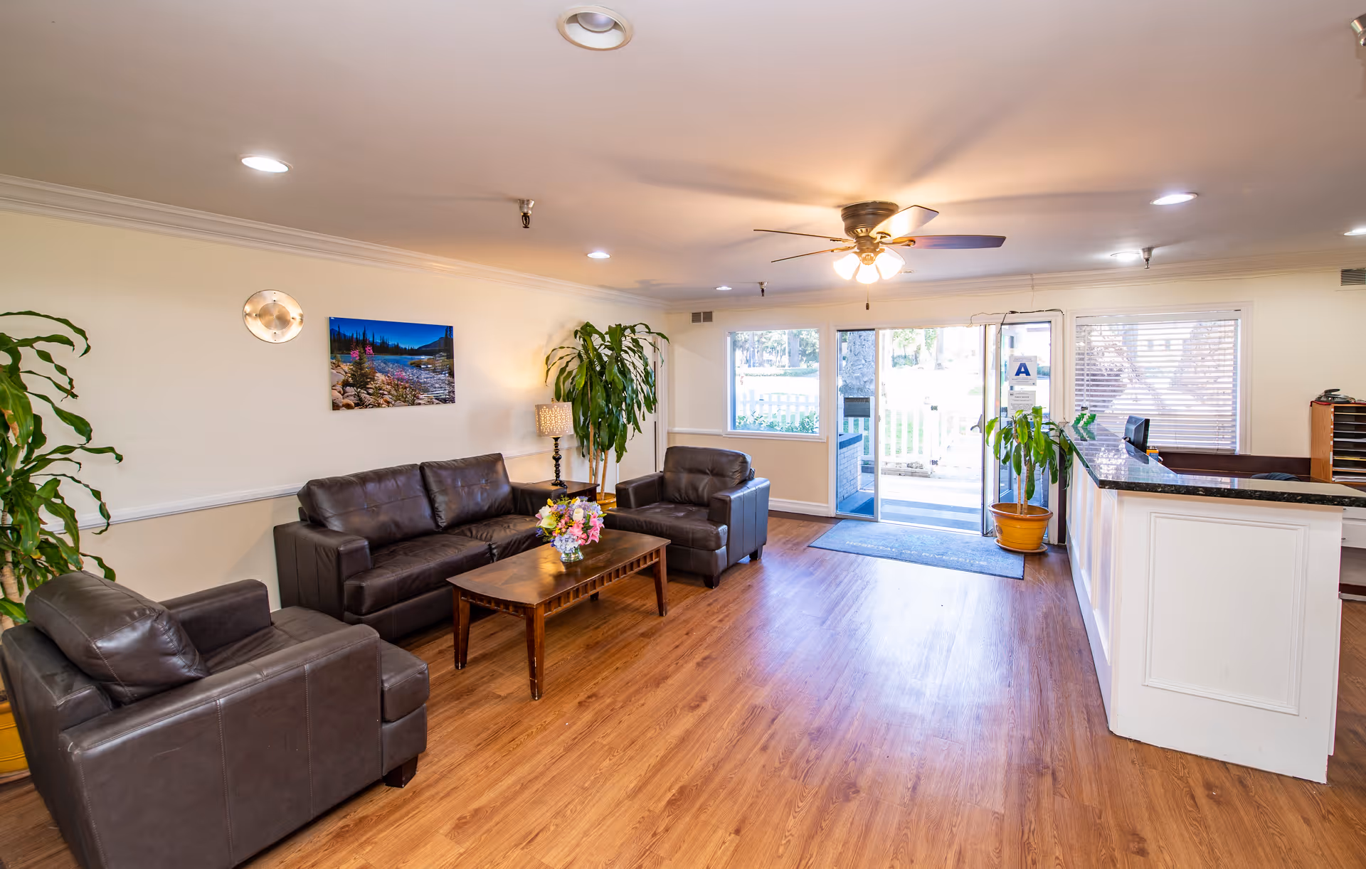 A bright and welcoming reception area with wooden flooring, a ceiling fan with lights, and large windows letting in natural light. The room features a dark brown leather sofa and two matching armchairs arranged around a wooden coffee table with a flower arrangement. There are several potted plants, a wall-mounted landscape picture, and a white reception desk with a granite countertop near the entrance door.