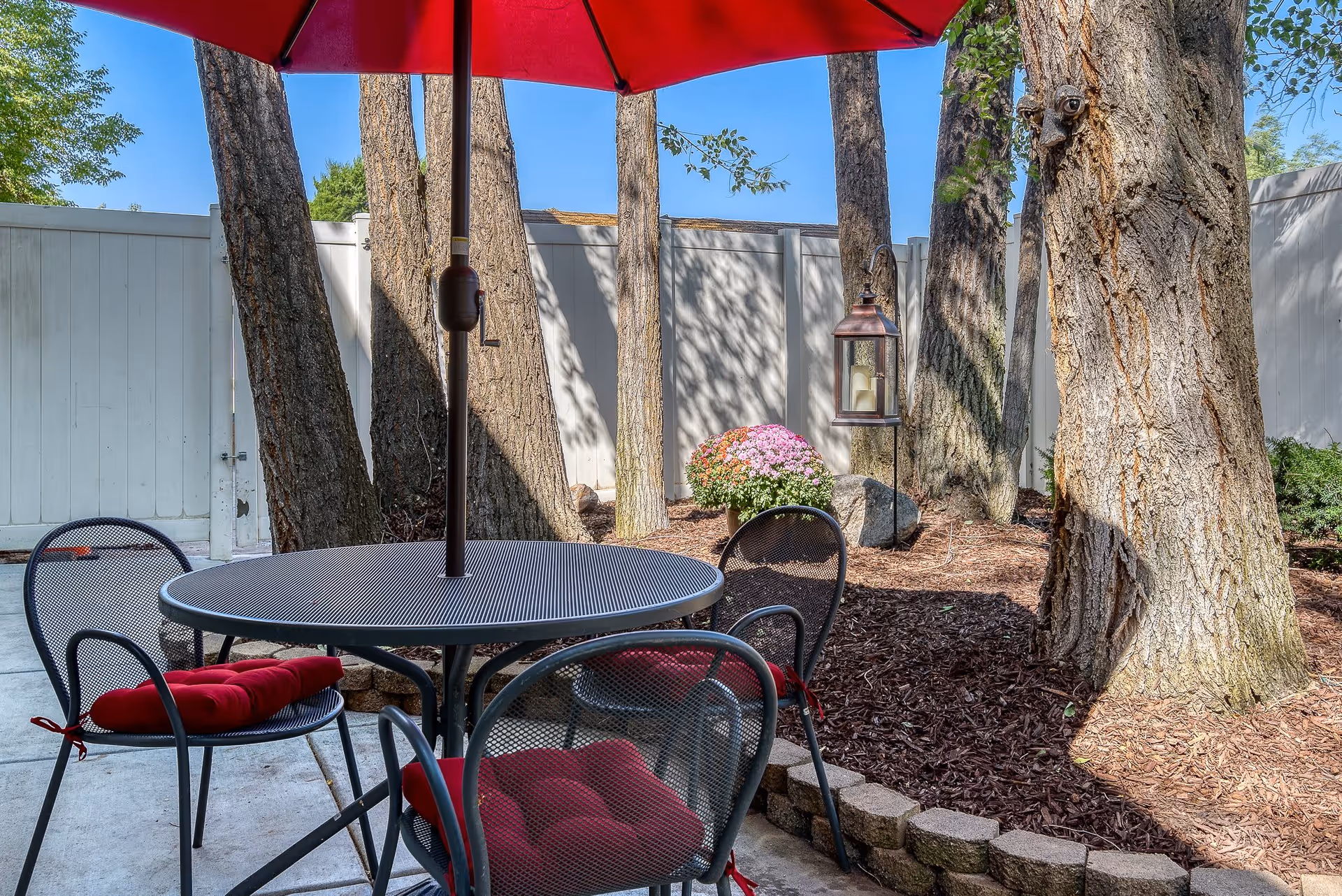 Outdoor patio area with a round metal table and four matching chairs with red cushions. A red umbrella is positioned over the table. The patio is surrounded by trees, a white fence, a flower pot with pink and orange flowers, and a hanging lantern with candles inside.