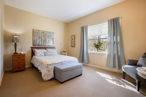 A bright and cozy bedroom with a double bed featuring white and light blue bedding. There is a wooden nightstand with a lamp and decorative items on the left side of the bed. A large window with light blue curtains lets in natural light, and a vase with red flowers is placed on the windowsill. A gray upholstered bench is at the foot of the bed, and a gray armchair with a small round table is positioned near the window. The walls are painted a soft beige color, and a framed artwork hangs above the bed.
