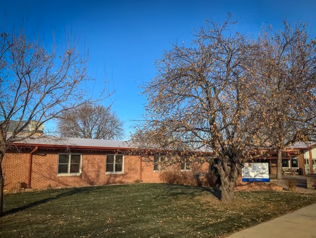 Exterior view of a single-story brick building with several windows, a large tree with sparse leaves in front, and a clear blue sky. There is a sidewalk and a grassy area surrounding the building.
