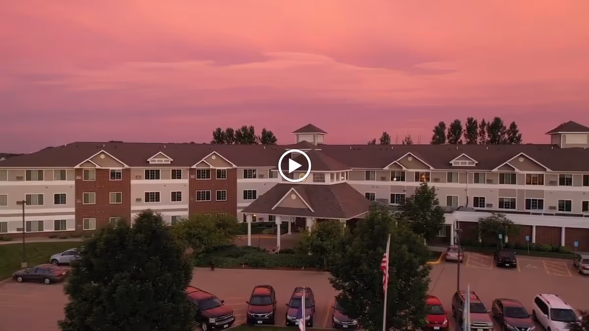 Exterior view of a large three-story senior living facility building named Summit Pointe during sunset with a pink and purple sky. The building has a covered entrance, multiple windows, and a parking lot with several cars parked. Trees and landscaping are visible around the building.