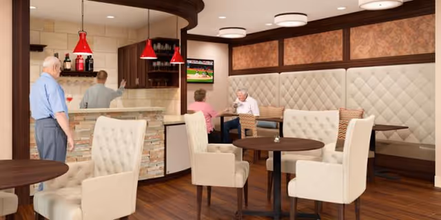 Interior view of a senior living facility lounge area with several beige upholstered chairs and round wooden tables. Two elderly people are seated and talking near a wall with tufted upholstery, while another elderly man stands near a counter with red pendant lights hanging above. A television is mounted on the wall showing a baseball game.