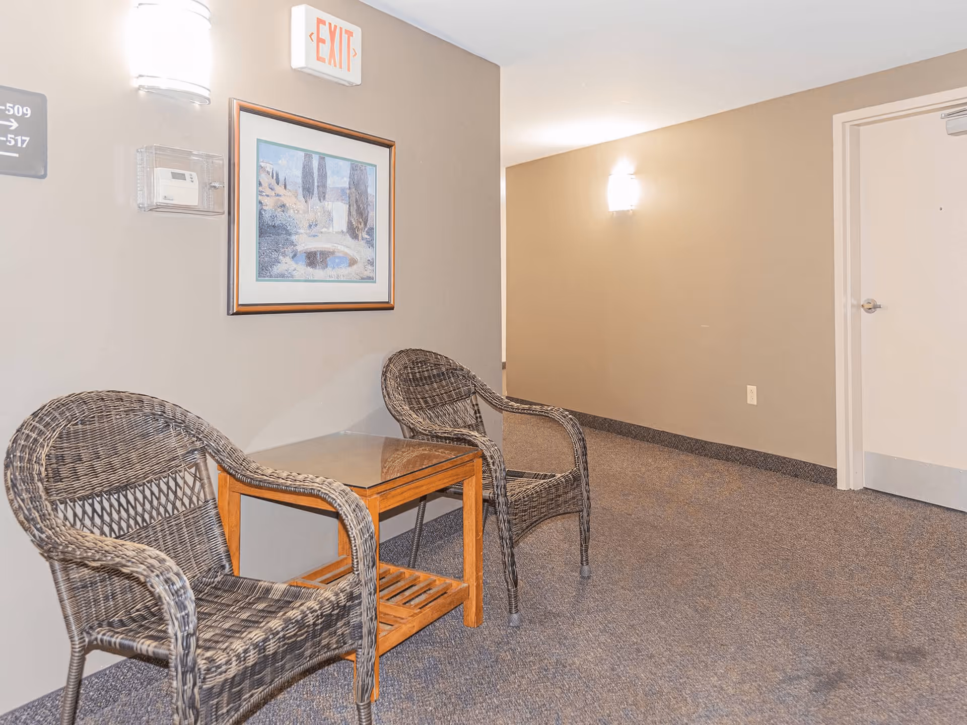 A hallway seating area with two wicker chairs and a wooden table with a glass top between them. The walls are beige with a framed landscape painting, wall-mounted lights, an exit sign, and a directional sign for rooms 509 to 517. The floor is carpeted and there is a closed white door at the end of the hallway.