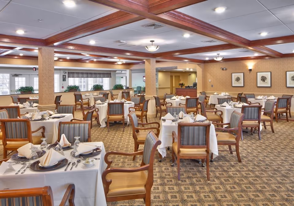 A spacious dining room with multiple tables covered in white tablecloths, each set with plates, napkins, glasses, and silverware. The room features wooden beams on the ceiling, patterned carpet, and framed artwork on the walls. Several wooden chairs with striped upholstery are arranged around the tables.