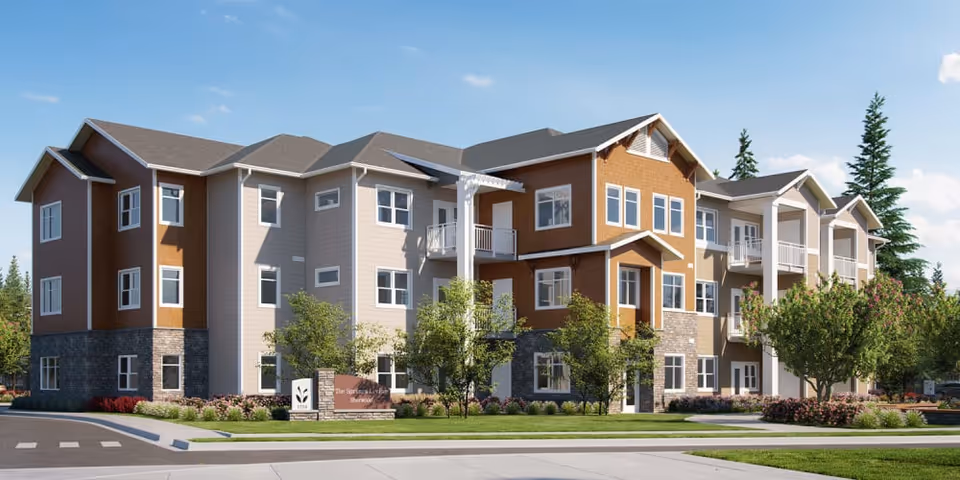 Three-story senior living building with balconies, landscaped lawns, and trees under a blue sky.
