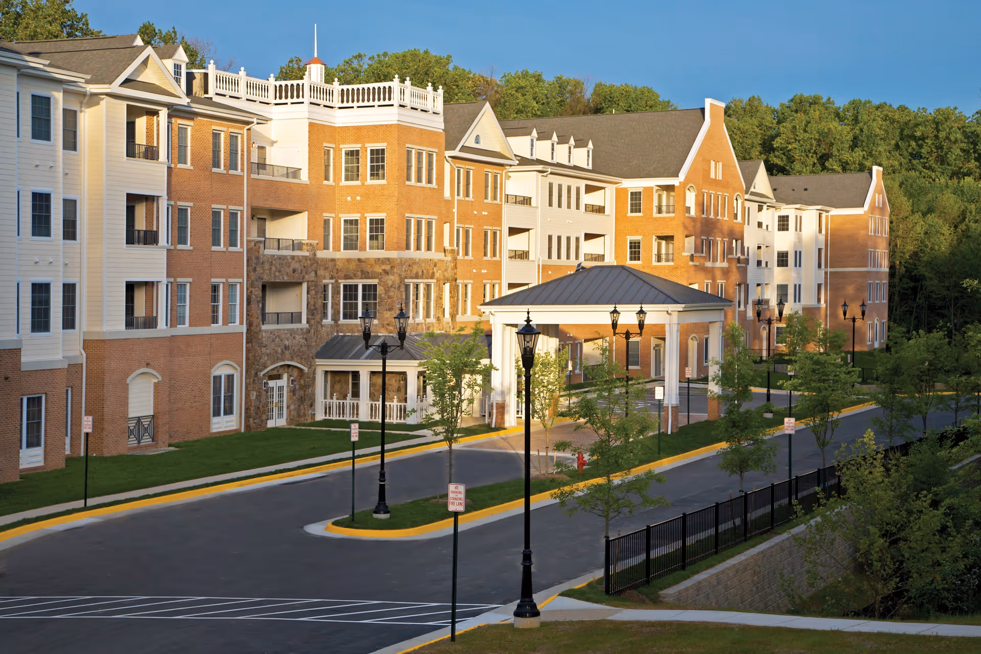 Exterior view of a large multi-story retirement community building with brick and siding facade, multiple windows, balconies, and a covered entrance driveway. The building is surrounded by trees and greenery under a clear blue sky.