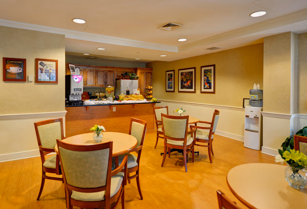 A cozy dining area in a senior living facility with round wooden tables and cushioned chairs. The room has warm yellow walls adorned with framed artwork. In the background, there is a kitchen counter with a coffee machine, a water dispenser, and a fruit stand. The floor is wooden, and small flower arrangements are placed on the tables.