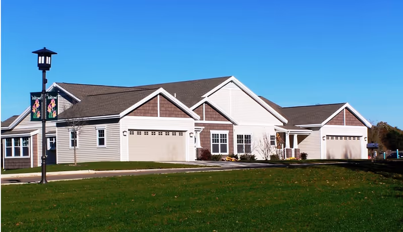 Single-story senior living building with attached garages, a manicured lawn, and a lamppost with banners under a clear blue sky.