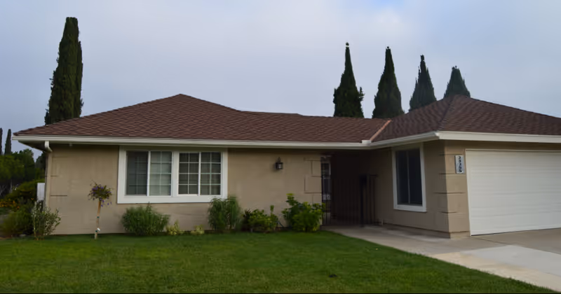 Single-story residential building with beige exterior walls, a brown shingle roof, white-framed windows, a garage door on the right, and a well-maintained green lawn with some shrubs and tall trees in the background under a cloudy sky.