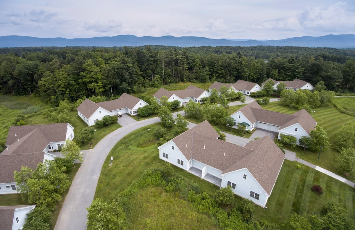 Aerial view of single-story white residential cottages arranged along a curved road surrounded by lawns and trees with distant mountains.