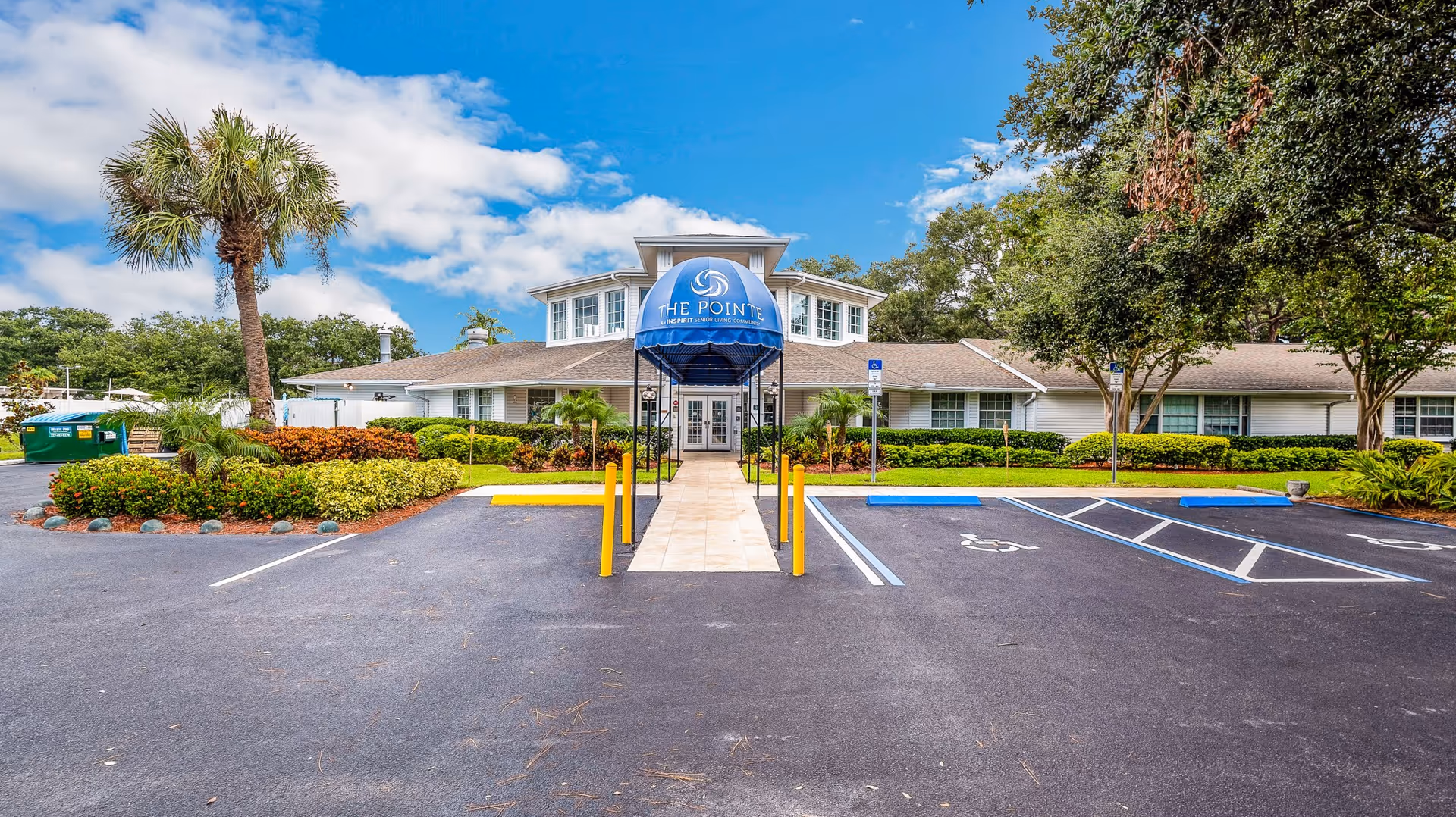 Front exterior view of The Pointe senior living community building with a blue canopy over the entrance, surrounded by landscaped greenery, palm trees, and a parking lot with handicap spaces.