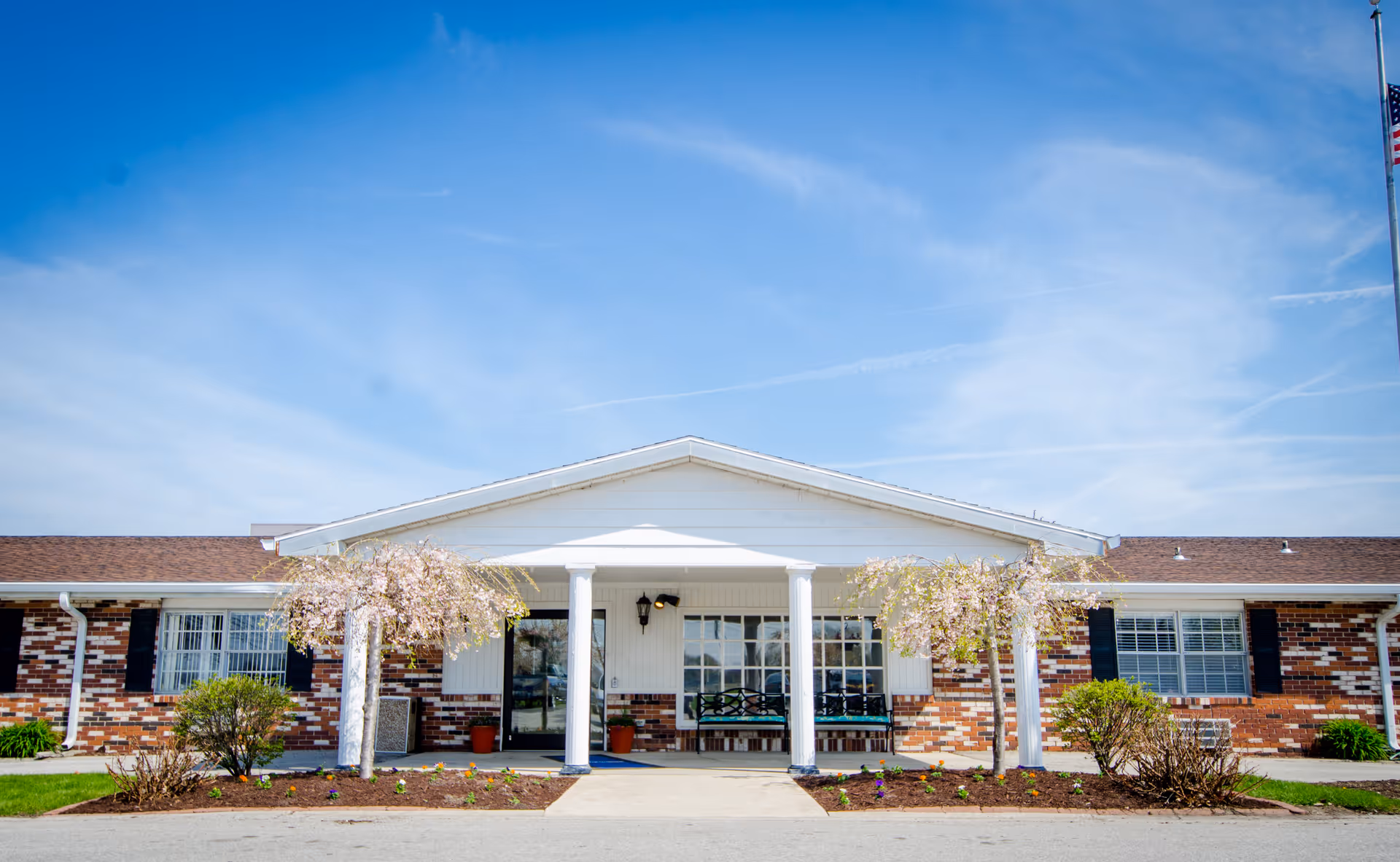 Front entrance of a single-story brick building with white columns, a covered portico, benches, and small flowering trees under a blue sky.