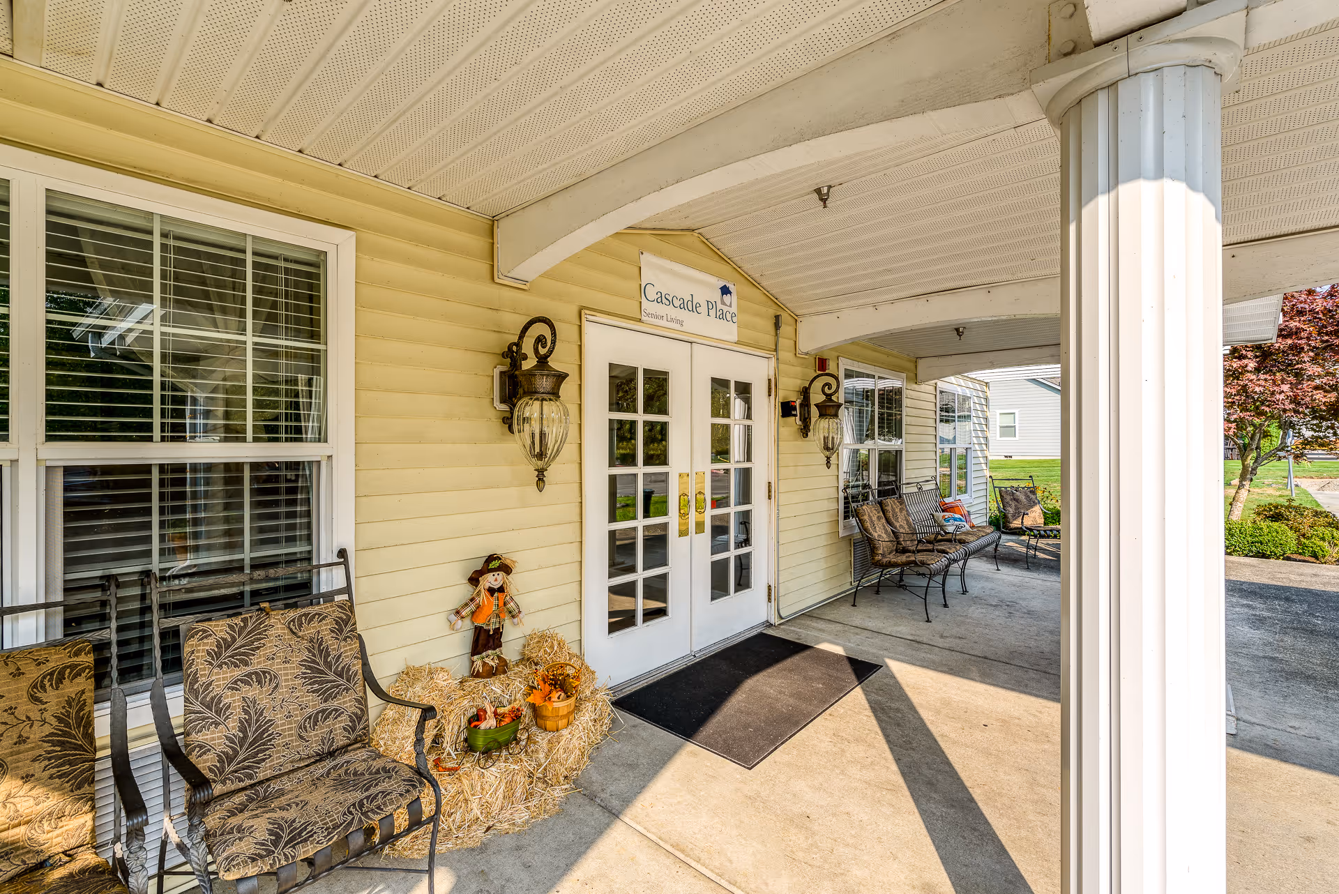 Covered front entrance of Cascade Place Assisted Living with double glass doors, seating, decorative fall display and a supporting column.
