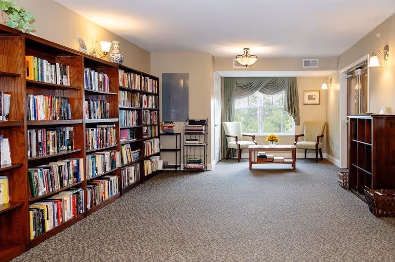 A quiet reading or sitting area in a senior living facility with wooden bookshelves filled with books on the left side, two armchairs and a small wooden table with a flower arrangement near a window with green curtains at the far end, and soft lighting from wall sconces and ceiling fixtures.