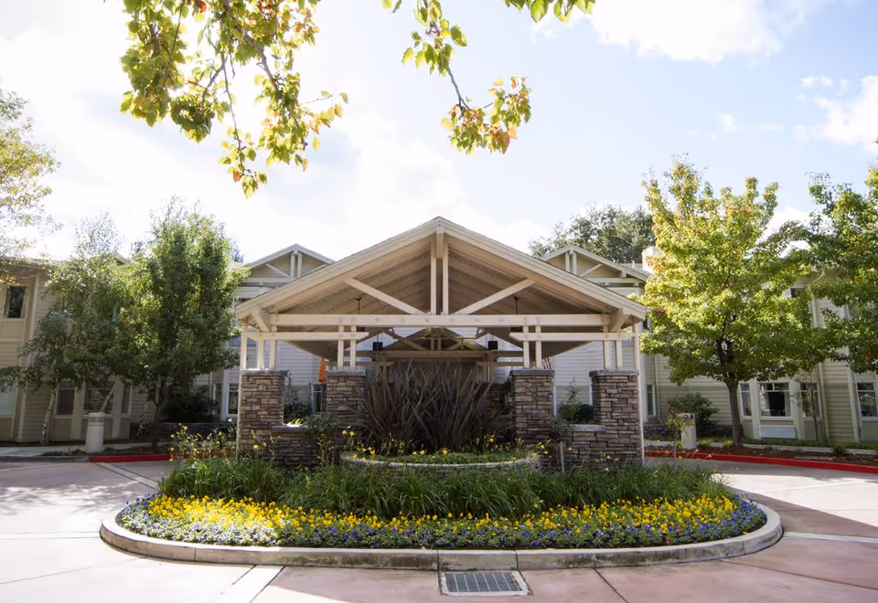 Entrance of a senior living facility with a covered driveway supported by stone pillars, surrounded by well-maintained landscaping including green trees and a circular flower bed with yellow and purple flowers.