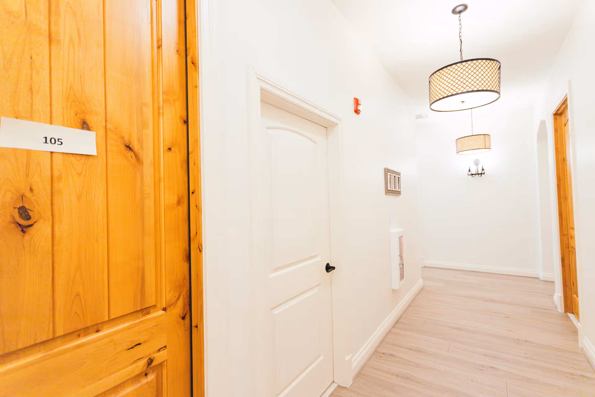 A bright hallway in a senior living facility with light wood flooring, white walls, and two hanging pendant lights with patterned shades. On the left, there is a wooden door labeled 105, and further down the hallway, there is a white door with a black handle. The hallway also features a wall-mounted light fixture and a fire safety box.