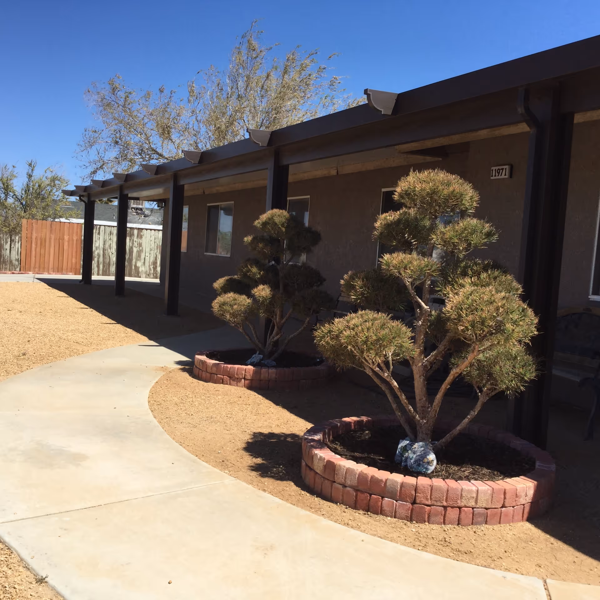 Outdoor view of Carmel Care Home showing a curved concrete walkway, two round brick-bordered garden beds with pruned bushes, a covered porch with dark wooden beams, a beige building wall with windows, and a clear blue sky.