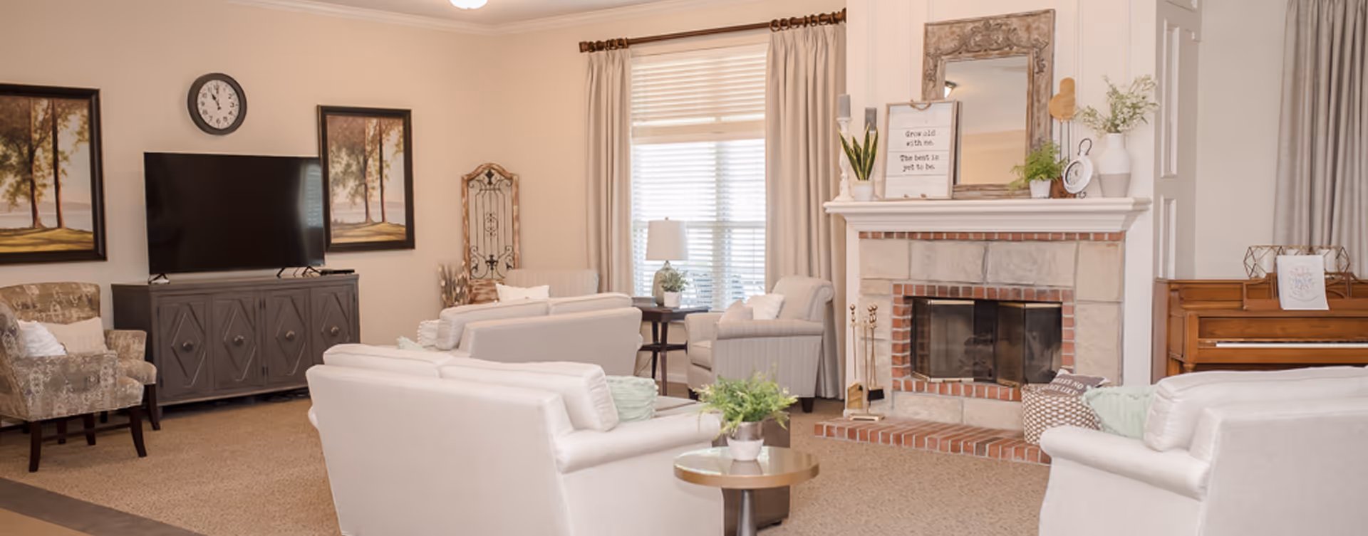 A cozy living room with beige walls and carpet, featuring a fireplace with a decorative mirror and plants on the mantel. The room has multiple armchairs and sofas arranged around a small round table with a plant. A flat-screen TV is mounted on a cabinet against the wall, flanked by two framed landscape paintings. A piano is visible on the right side near a window with curtains.
