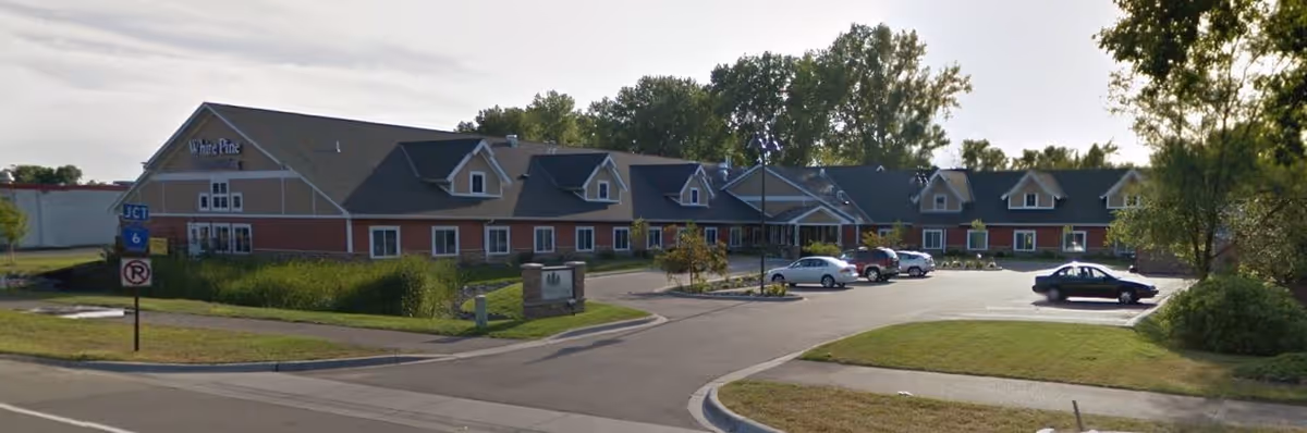 Exterior view of White Pine Advanced Assisted Living and Memory Care facility in Fridley, showing a large building with multiple dormer windows, a parking lot with several cars, and surrounding greenery under a partly cloudy sky.