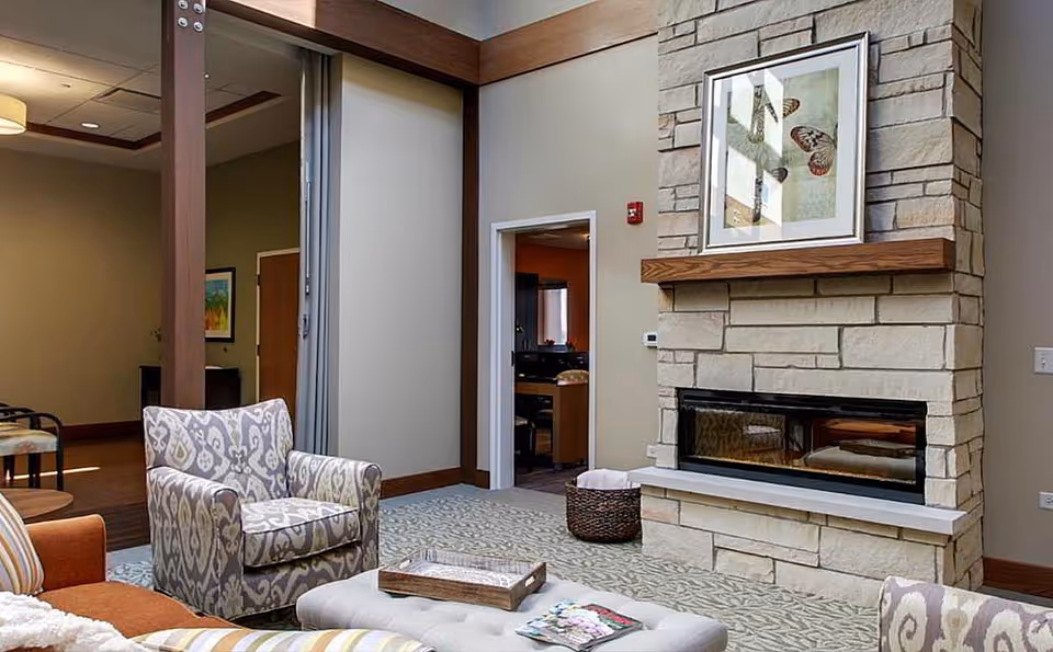 A cozy living room area in a senior living facility featuring a stone fireplace with a wooden mantel and framed butterfly artwork above it. The room has patterned armchairs, a cushioned ottoman with a tray and magazines, and neutral-toned walls with wooden trim. An open doorway leads to another room with a desk and chair.