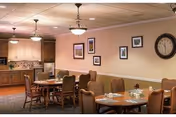 Interior view of a dining room with several wooden tables and cushioned chairs. The tables are set with placemats, napkins, and utensils. The room has beige walls adorned with framed pictures and a large clock. Ceiling lights hang above, and a kitchen area with cabinets and appliances is visible in the background.